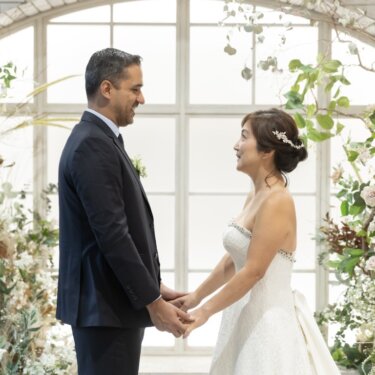 洋装に着替えた新郎とウェディングドレスの花嫁が、花のアーチ窓のセットで手を取り合う正面の結婚写真