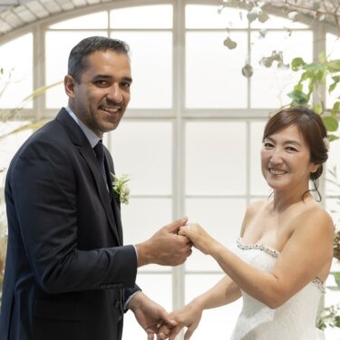 アーチ窓と花のガーデンセットで手を取り合い、カメラに笑顔を向ける新郎新婦のスタジオ前撮り