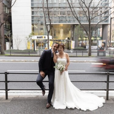 街並みを望む歩道の柵にもたれ、頬に手を添えて微笑む新郎新婦。都会的な横浜の街並みの前撮り