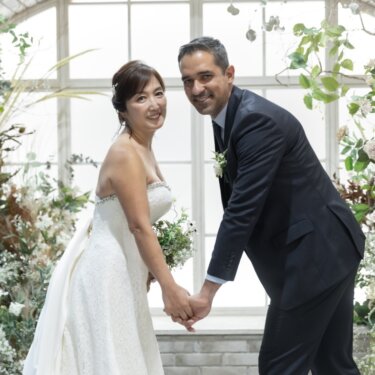 アーチ窓と花々のガーデンセットで、手をつないで前かがみになりカメラ目線で笑う新郎新婦のスタジオフォト