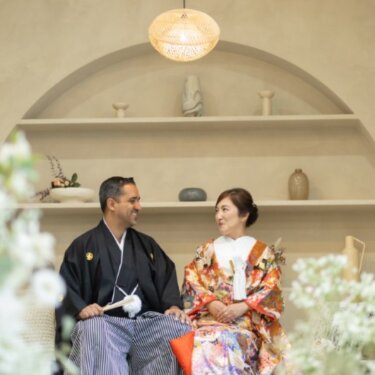 アーチ型のニッチと竹の照明、陶器の飾り棚の前に座って見つめ合う和装の新郎新婦のスタジオフォト