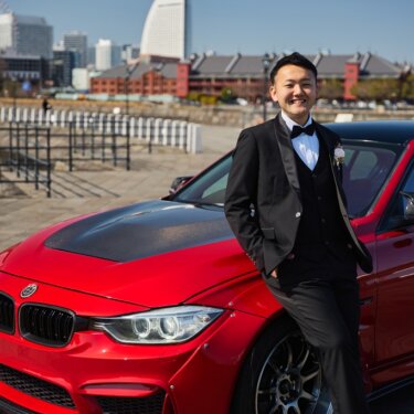 赤レンガ倉庫とランドマークタワーを望み、赤い愛車に寄りかかるタキシードの新郎のソロの前撮り