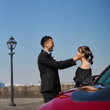 港のガス灯と赤い愛車のそばで、花嫁のほおをやさしくつまむ新郎。遊び心あるウェディングフォト
