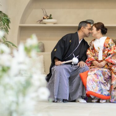 白い花を前景にぼかし、アーチの棚のセットで鼻先を寄せ合う新郎新婦の和装スタジオフォト