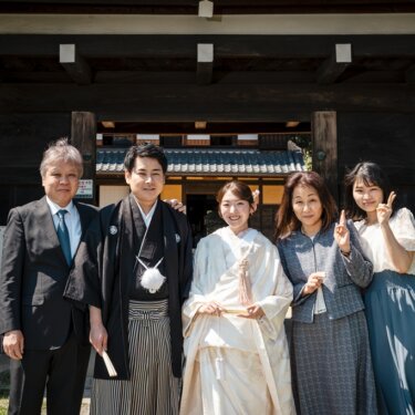 茅葺屋根の長屋門の前で、両家の家族と笑顔で並ぶ白無垢の花嫁と新郎。前撮りで撮る家族写真