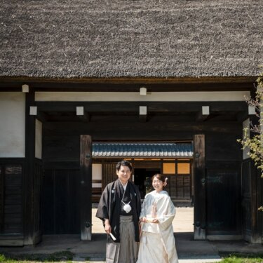 横溝屋敷の茅葺屋根の長屋門の前に並んで立つ、紋付袴の新郎と白無垢の花嫁の和装前撮り