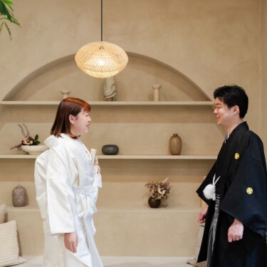 壺や置物が飾られた洋風の壁棚を背に、見つめ合う白無垢の花嫁と黒紋付の新郎の和装スタジオフォト