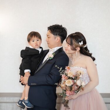 壁灯のある白い壁の前で、息子を抱く新郎とほほえむカラードレスの花嫁の家族写真のスタジオフォト