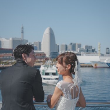 みなとみらいのビル群を望む大さん橋の柵際で、振り返って笑う花嫁と新郎。バックスタイルのドレスが映える前撮り