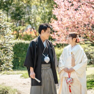 濃いピンクの桜の前で見つめ合う紋付袴の新郎と白無垢の花嫁。手毬の小物が映える和装前撮り
