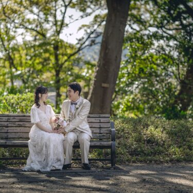 緑の木々に囲まれた木のベンチに並んで座り、見つめ合う新郎新婦。公園で撮るナチュラルなウェディングフォト