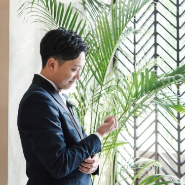 観葉植物とアイアンの窓を背に、袖のカフスを整えるネイビーのタキシードの新郎のソロショットのスタジオフォト