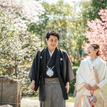 濃いピンクの桜を背に、扇子と手毬を手に笑い合う白無垢の花嫁と紋付袴の新郎の和装前撮り