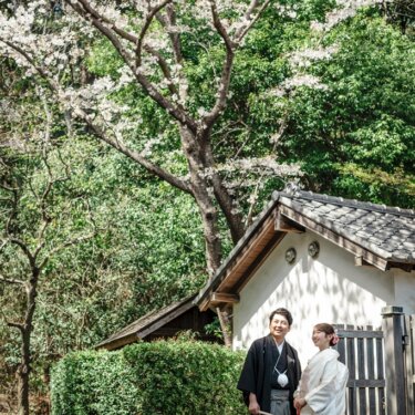 横溝屋敷の桜の咲く庭で、白壁の土蔵のそばに立つ紋付袴の新郎と白無垢の花嫁の和装フォトウェディング