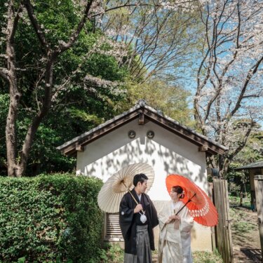 白壁の土蔵の前で、白と朱赤の和傘を差して向かい合う新郎新婦。横溝屋敷の庭で撮るロケーションフォト