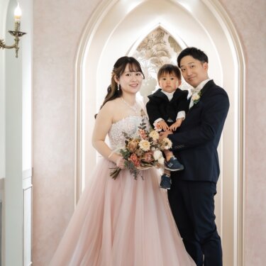 尖塔アーチのセットで息子を抱いた新郎と、ピンクのカラードレスの花嫁が並ぶ家族写真の前撮り