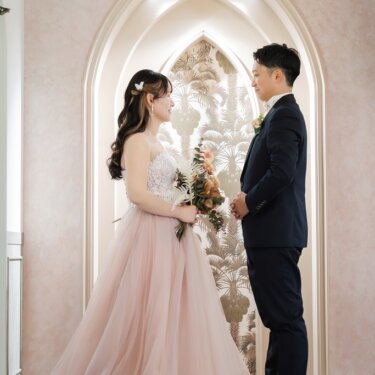 尖塔アーチのセットで見つめ合う新郎とピンクのチュールのカラードレスの花嫁。トレーンが広がる結婚写真