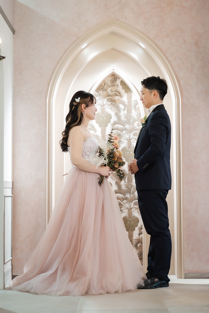 尖塔アーチのセットで見つめ合う新郎とピンクのチュールのカラードレスの花嫁。トレーンが広がる結婚写真