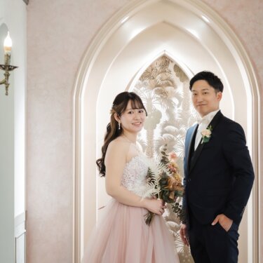 ゴシック調のアーチのセットに並んで立ち、カメラを見つめる新郎とピンクのカラードレスの花嫁のスタジオフォト