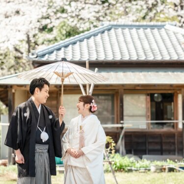 横溝住宅にて、白い和傘の下で笑い合う紋付袴の新郎と白無垢の花嫁。満開の桜と古民家が背景の和装フォトウェディング