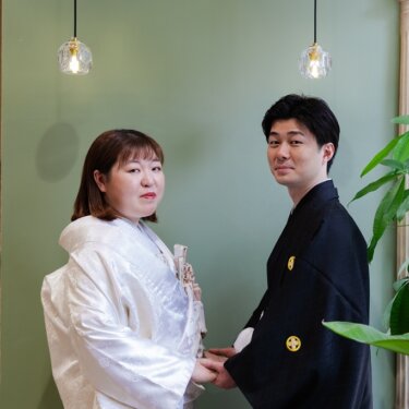 淡いグリーンの壁とペンダントライトを背に、手をつないでカメラを見つめる和装の新郎新婦のスタジオフォト
