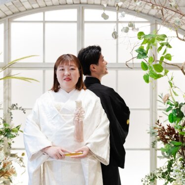 大きなアーチ窓とバラの装飾を背景に、背中合わせで立つ白無垢の花嫁と紋付袴の新郎の和装スタジオフォト