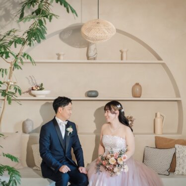 陶器の飾り棚とラタンの照明を背に、クッションのあるベンチに座って見つめ合う新郎新婦のスタジオフォト