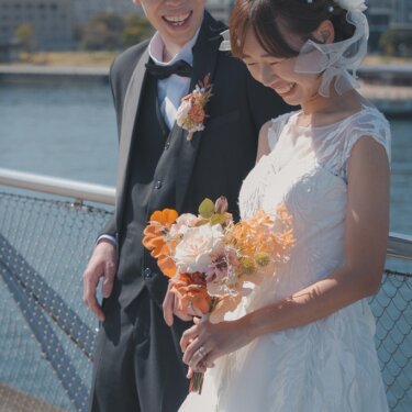 寄り添ってブーケを見つめ、大笑いする新郎新婦。港の景色をぼかした自然体の結婚写真