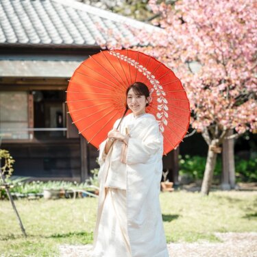 桜と古民家を背に、朱赤の和傘を差してほほえむ白無垢の花嫁のソロショット。横溝屋敷で撮る春の和装前撮り