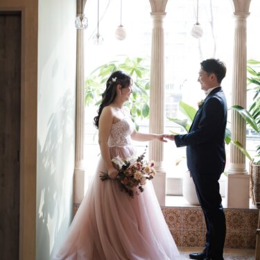 大きなアーチ窓と観葉植物、円柱の並ぶスタジオで、手を差し出して見つめ合う新郎新婦の全身のスタジオフォト