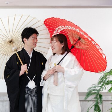 白と朱赤の和傘を差して見つめ合う、紋付袴の新郎と白無垢の花嫁。グリーンが映えるスタジオの和装フォトウェディング