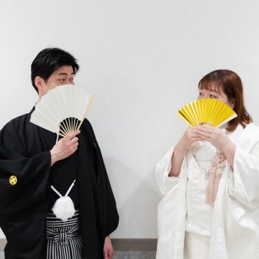 白と金の扇子で口元を隠し、いたずらっぽく見合う紋付袴の新郎と白無垢の花嫁。スタジオで撮る和装前撮り