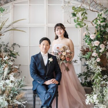 花のアーチと窓のセットで、椅子に座る新郎とそばに立つピンクのカラードレスの花嫁の結婚写真
