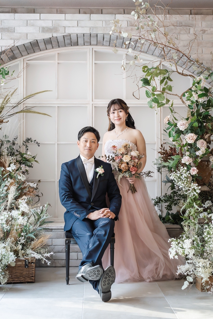 花のアーチと窓のセットで、椅子に座る新郎とそばに立つピンクのカラードレスの花嫁の結婚写真