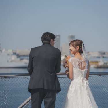 柵際で振り返ってほほえむ花嫁と、港を眺める新郎の後ろ姿。背中のリボンがかわいいウェディングドレスの前撮り