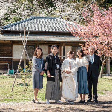 横溝住宅にて、桜が咲く古民家の庭で、家族と並んで立つ新郎新婦を引きで写した家族写真の和装フォトウェディング