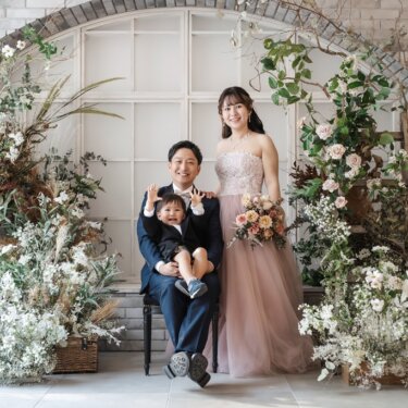 花のアーチを背に、息子が両手を上げる家族3人の記念写真。カラードレスで撮るフォトウェディング
