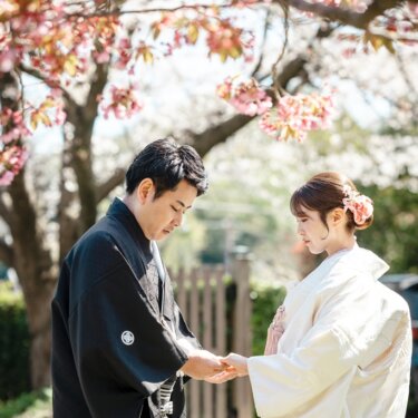 桜の木の下で白無垢の花嫁の手を取り、指輪を見つめる紋付袴の新郎。春らしい和装の結婚写真