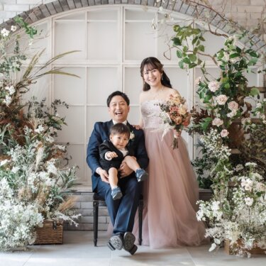 花のアーチのセットで、椅子に座る新郎が息子を抱き、ピンクのドレスの花嫁が寄り添う家族写真のスタジオフォト