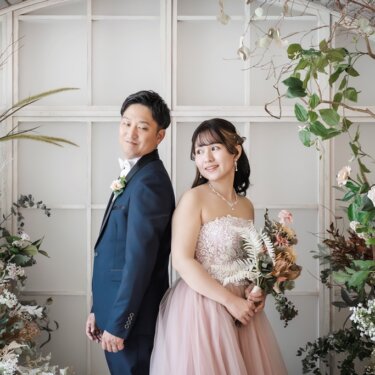 花のアーチを背に背中合わせで振り返る、ネイビーのタキシードの新郎とピンクのカラードレスの花嫁のスタジオフォト