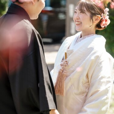 桜の花を前景にぼかし、新郎と見つめ合ってほほえむ白無垢の花嫁のアップの和装前撮り
