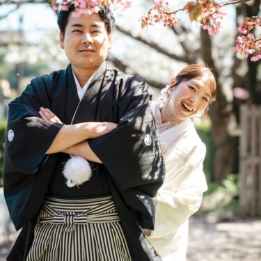 腕を組んで正面を向く紋付袴の新郎の後ろで、笑顔をのぞかせる白無垢の花嫁。桜の季節の和装フォトウェディング