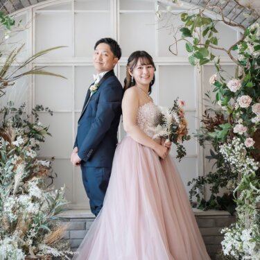 ドライフラワーの装花とアーチ窓のセットで、背中合わせに立つ新郎とピンクのカラードレスの花嫁の前撮り