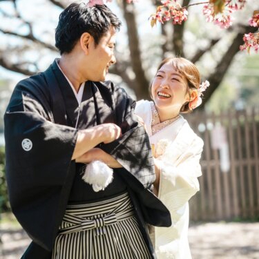 桜の下で腕を組む新郎と、後ろからのぞき込んで笑う白無垢の花嫁。遊び心あるポーズの和装前撮り