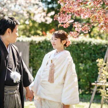 桜の枝の下で手をつないで見つめ合う新郎新婦。ピンクの髪飾りが映える白無垢の結婚写真