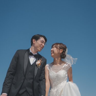 青空を大きく入れたローアングルで、見つめ合って笑う新郎新婦。タキシードとウェディングドレスの結婚写真