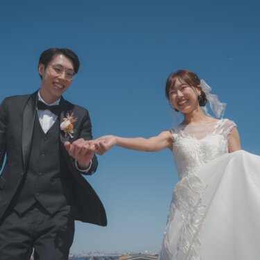 真っ青な空の下、ドレスの裾を持ち上げて新郎と手をつなぐ花嫁。躍動感あるローアングルの前撮り