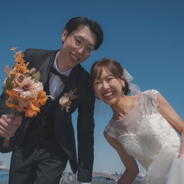 青空を背景に身を寄せ合って大笑いする新郎新婦のアップ。オレンジのブーケが華やかなフォトウェディング