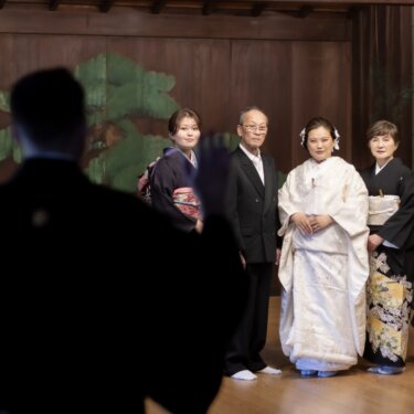 松の鏡板の前に並ぶ白無垢の花嫁と家族を、新郎の後ろ姿越しに写した家族写真のフォトウェディング