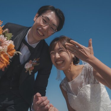 片手を掲げてはしゃぐ花嫁と、ブーケを持って笑う新郎を青空とともに写した大さん橋の結婚写真
